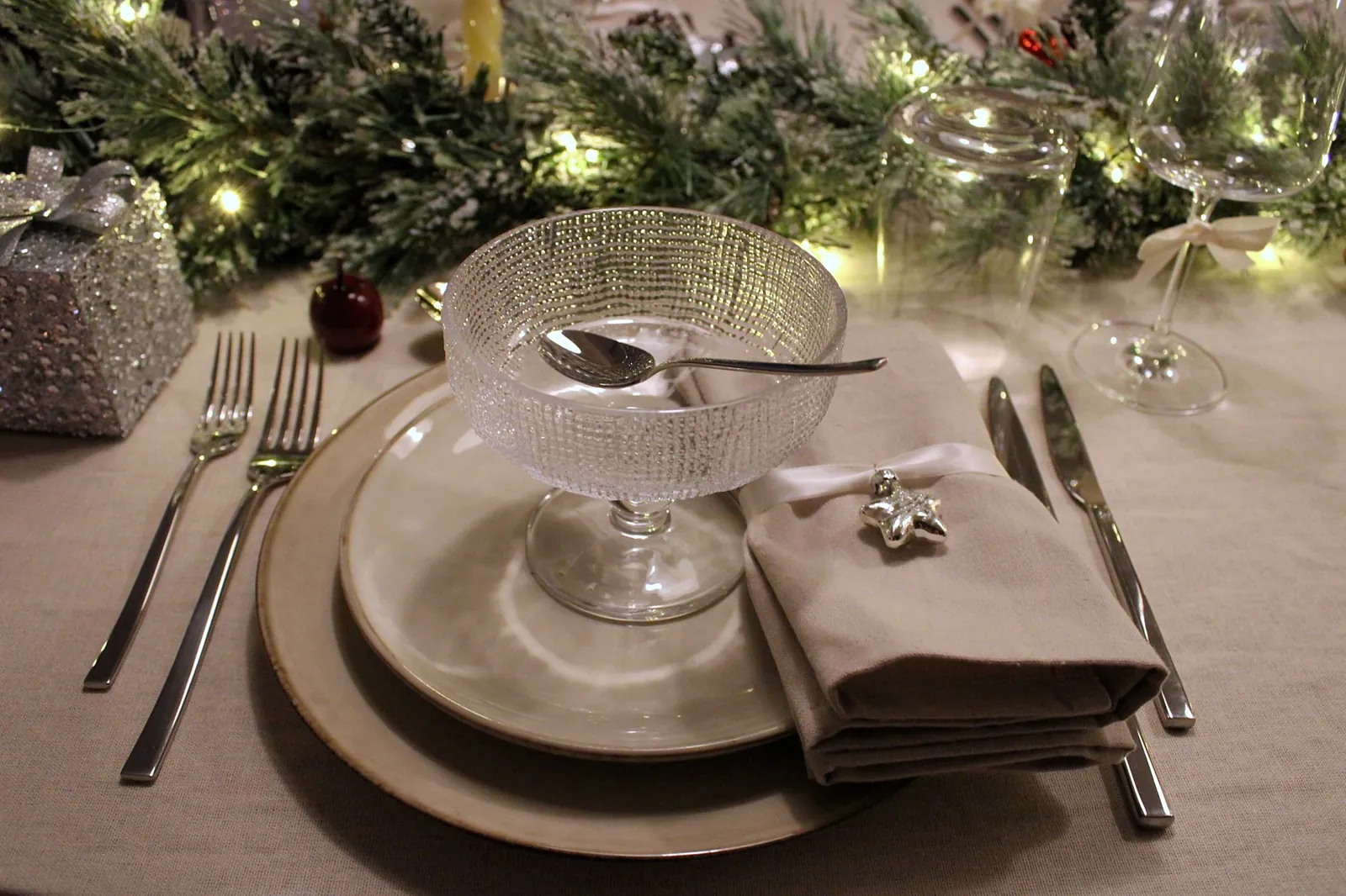 Couvert in de Winter Glow-collectie: dessertcoupe op het bord, katoenen servet met lint en zilveren ster, dennenslinger met lichtjes op de achtergrond